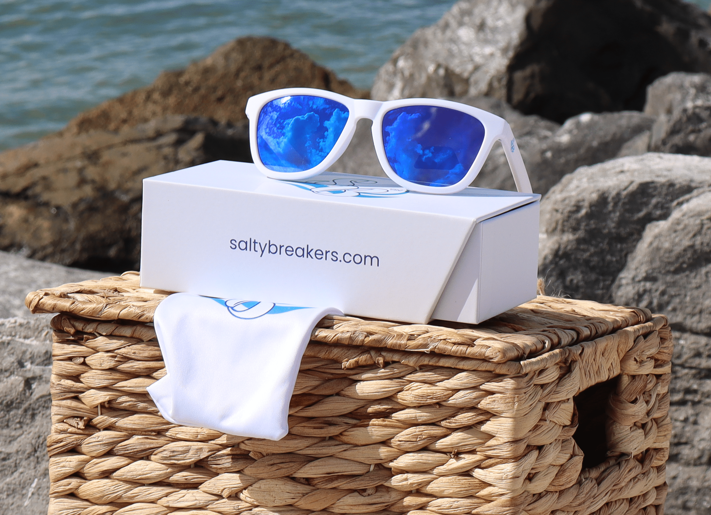 Salty Breakers™ Malibu polarized sunglasses in matte white with reflective blue mirror lenses resting on a Seafoam White presentation box and branded microfiber storage pouch atop a woven basket against sea jetty rocks.